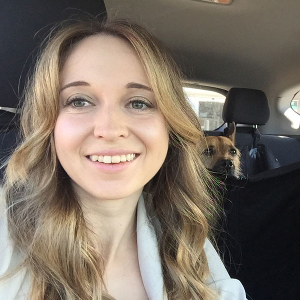 A photo of Jamie Hill-Borton smiling at the camera in her car with her dog in the backseat of the background