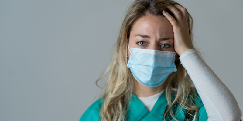 Woman in teal scrubs wearing a blue surgical mask, hand on head in distress.