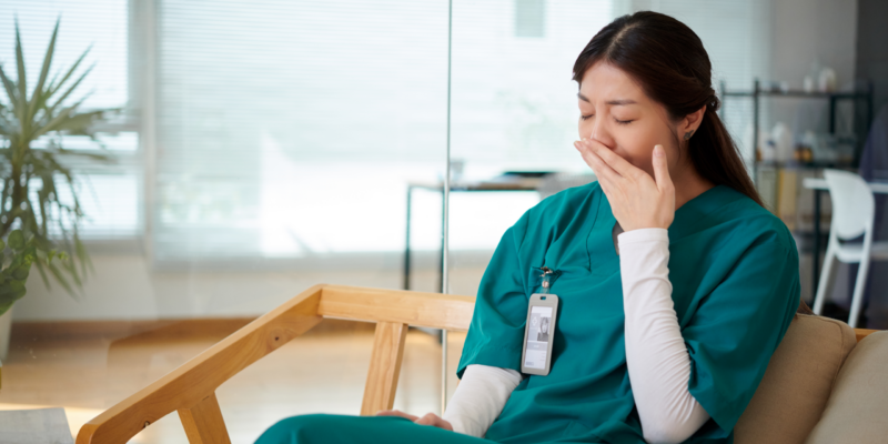 Nurse in teal scrubs sits in a clinic, covering her mouth with a hand as if yawning or coughing.